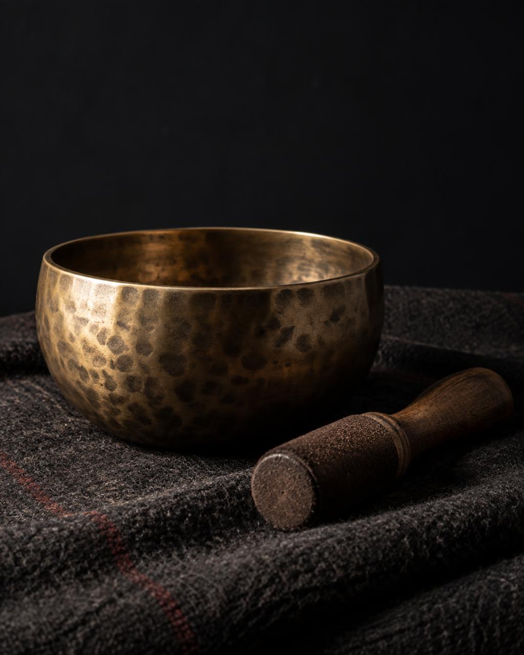 A hand-hammered brass singing bowl resting on a dark woven blanket, with a suede-wrapped mallet beside it
